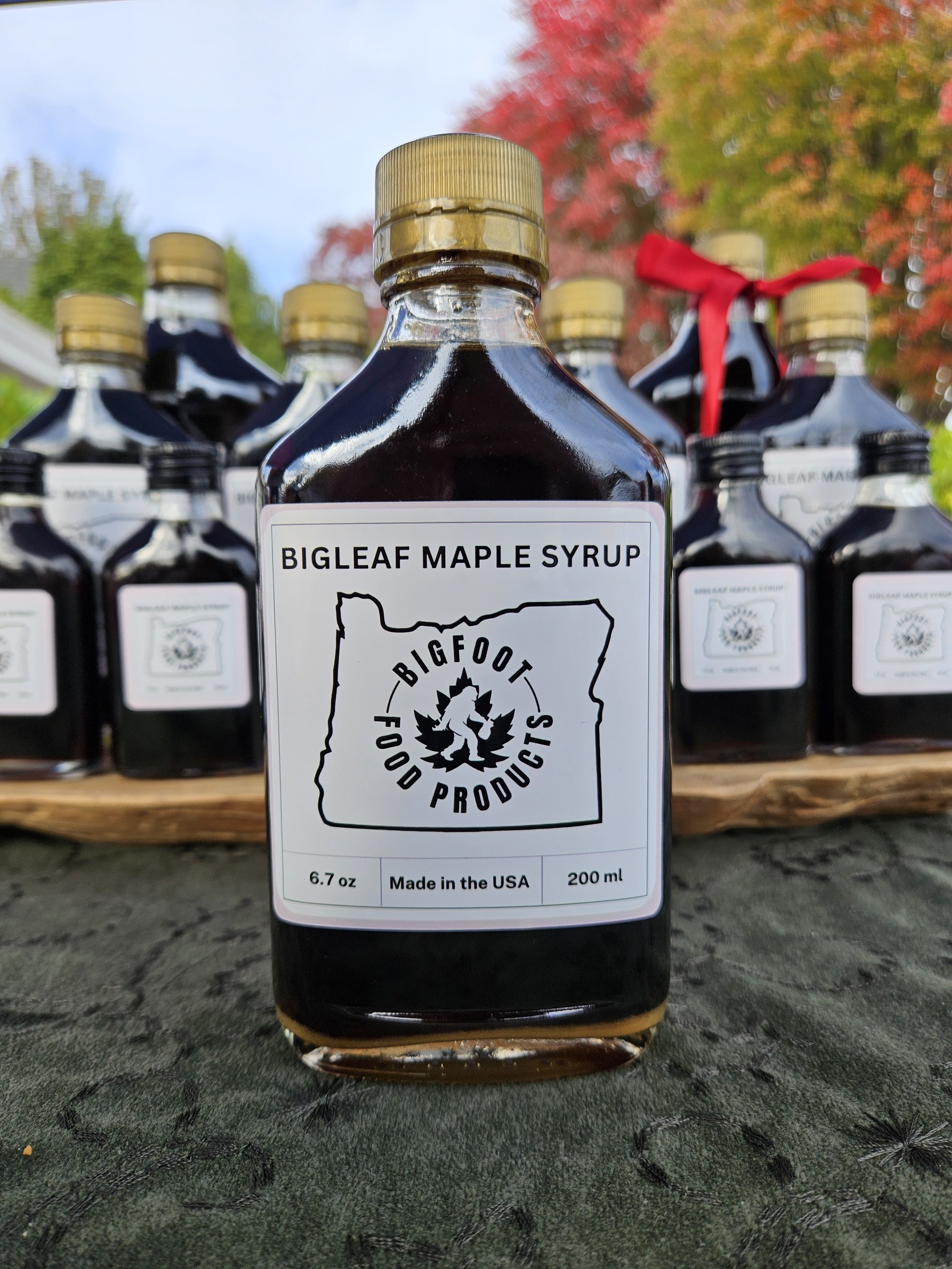 Bigfoot Bigleaf Maple Syrup 200ml bottle with gold cap in front of a row of bottles, fall leaves behind